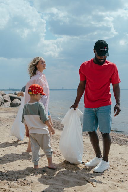 A man in a red t-shirt, denim shorts, and a black cap is standing on a sandy beach, holding a large white trash bag in his left hand while appearing to collect litter. To his right, a young boy wearin