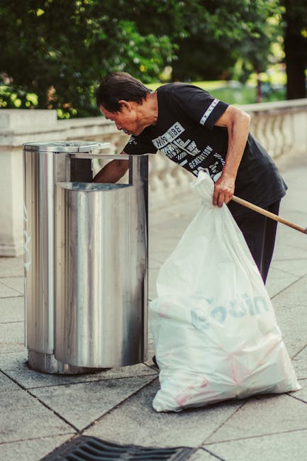 A black wheeled rubbish bin labeled 'St. John's' positioned on the edge of a sidewalk during nighttime. The bin's lid is open, revealing scattered cardboard boxes and miscellaneous waste materials ins